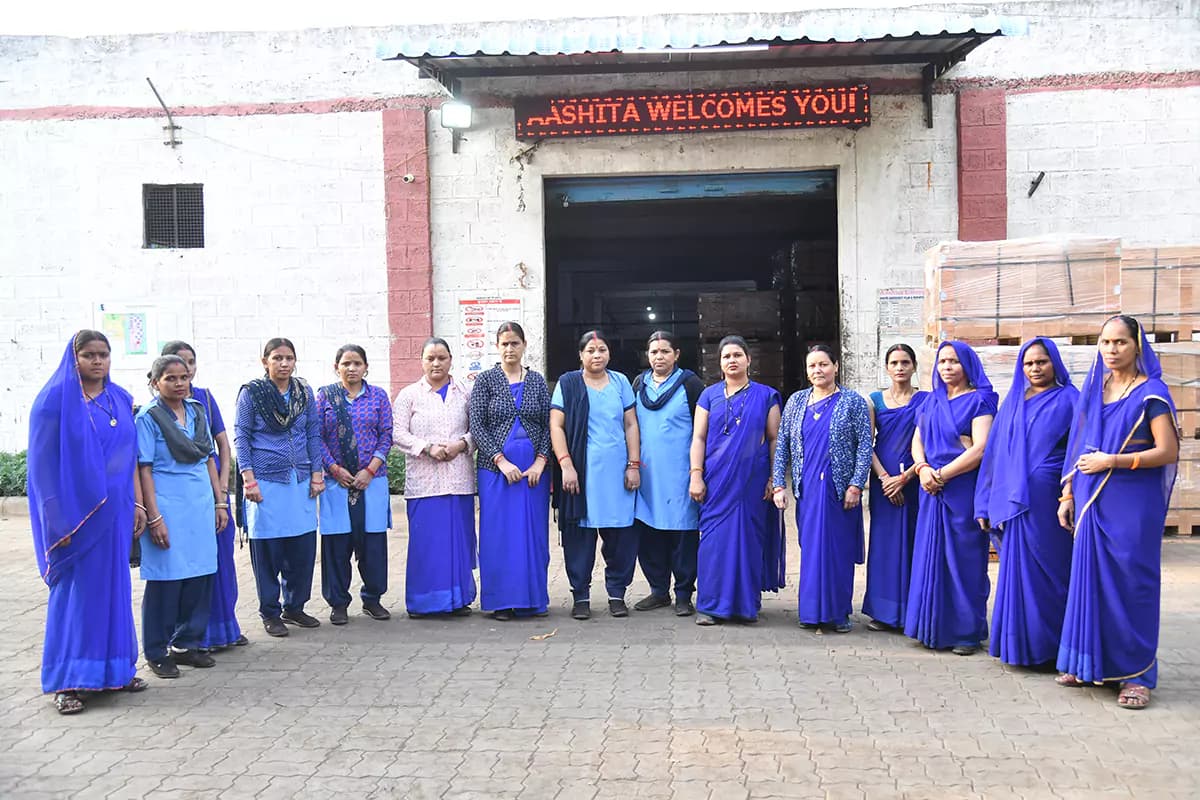 Members of the production team outside the plant entrance beneath an Aashita Welcomes You sign